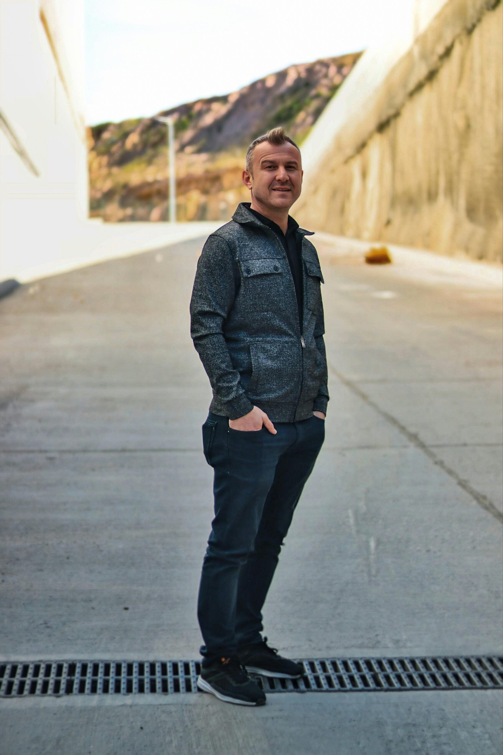 Man in casual outfit smiling, standing on a sunny day outdoors, showcasing urban and natural blend.