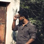 A man adjusting his sunglasses stands stylishly by a rusty door, surrounded by nature.