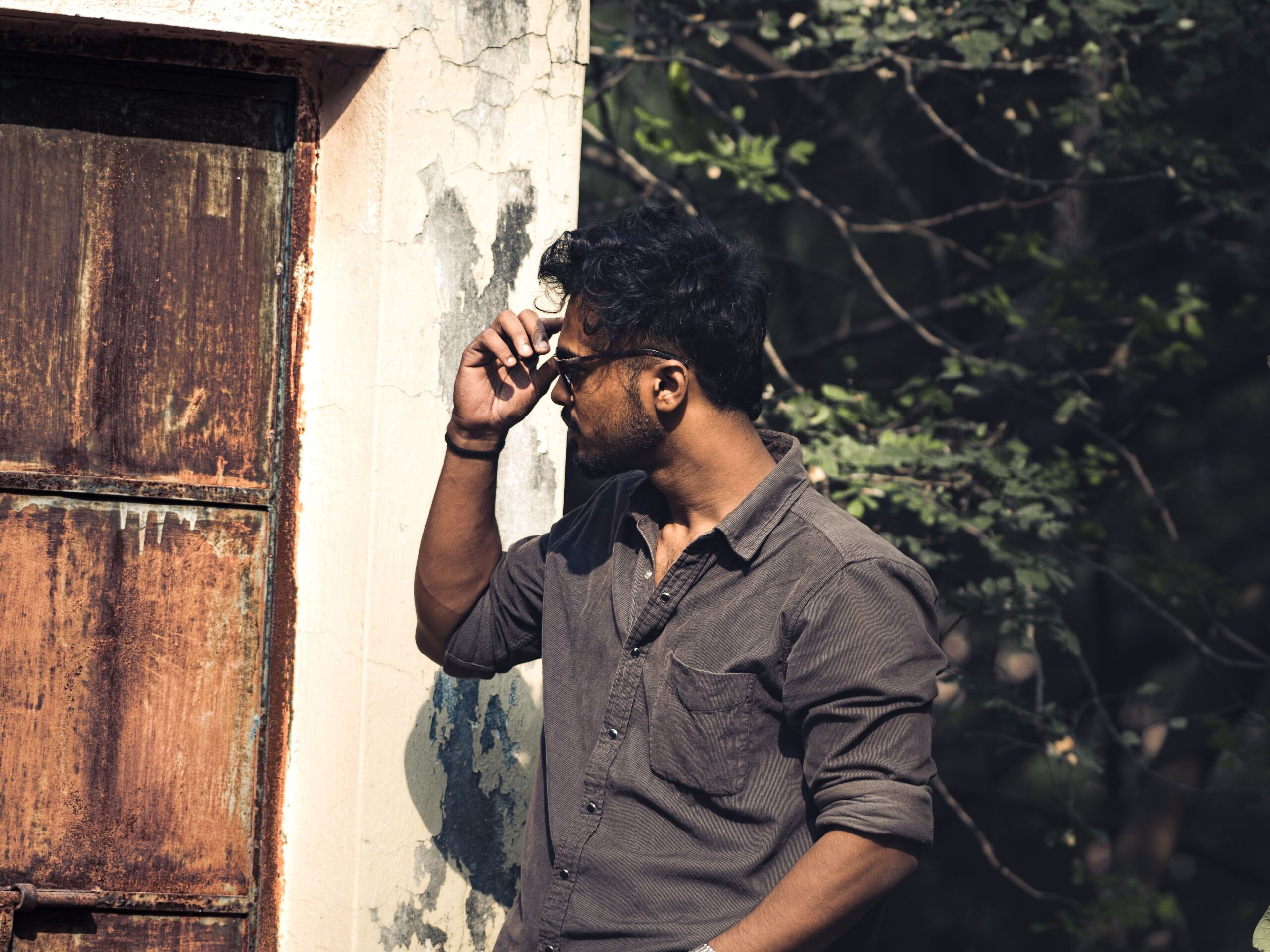 A man adjusting his sunglasses stands stylishly by a rusty door, surrounded by nature.