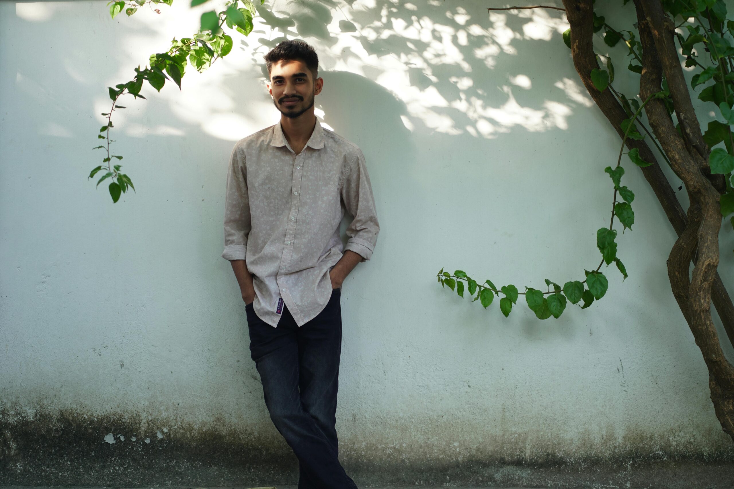 A young man casually leaning against a white wall with greenery.